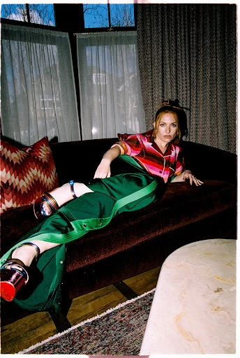 Adult woman reclining on a sofa in a photo studio setting, wearing a striped shirt and green pants with heels, with a stylish interior.