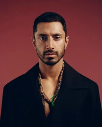 Portrait of a man with short dark hair and a beard wearing a black jacket and layered necklaces against a solid dark red background