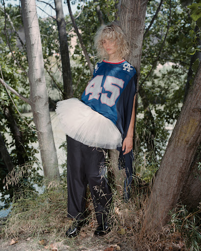 Blonde woman in a blue sports jersey and white tutu poses in a wooded area with trees and a body of water in the background.