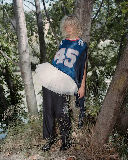 Blonde woman in a blue sports jersey and white tutu poses in a wooded area with trees and a body of water in the background.