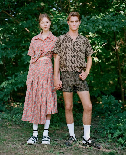 A young couple, a male and female, stand in a forest clearing wearing matching outfits with designer patterns. They are holding hands.