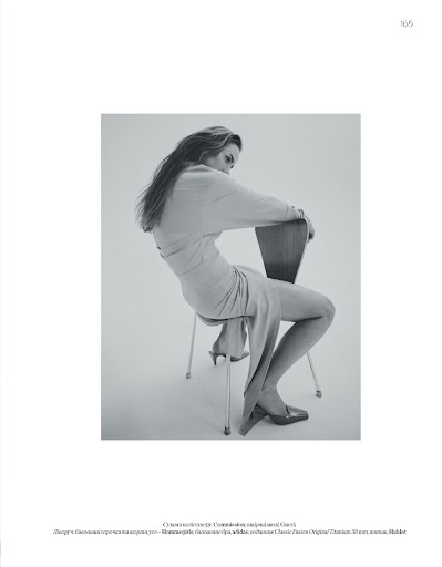 Monochrome fashion portrait of a woman in a long dress seated backward on a wooden chair against a white background.