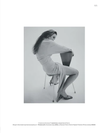 Monochrome fashion portrait of a woman in a long dress seated backward on a wooden chair against a white background.