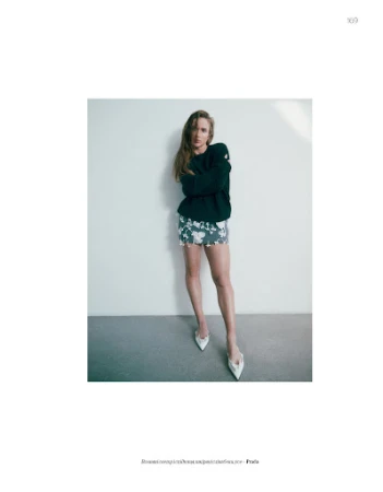 Full-length portrait of a woman in a dark sweater and patterned skirt standing against a pale seamless studio background.