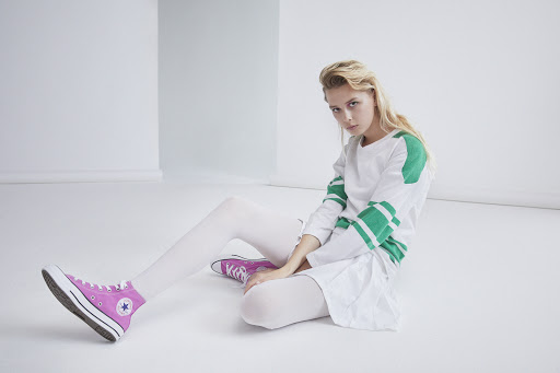 Blonde woman in a white top and skirt sitting on a white floor with pink sneakers in a minimalist studio setting.
