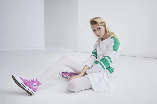 Blonde woman in a white top and skirt sitting on a white floor with pink sneakers in a minimalist studio setting.