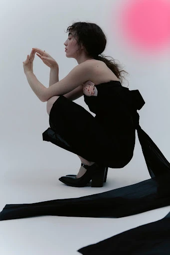 A woman with dark hair poses in a black dress with a ribbon and black heeled shoes against a gradient pink and white background.
