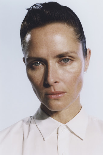 Headshot portrait of an adult woman with slicked-back dark hair and a white collared shirt against a neutral light blue background.