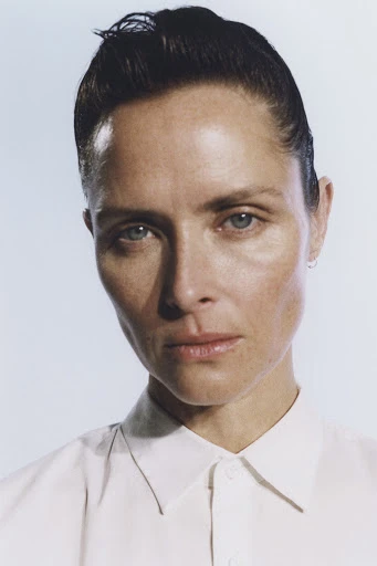 Headshot portrait of an adult woman with slicked-back dark hair and a white collared shirt against a neutral light blue background.