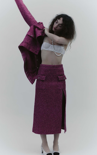 A woman in a stylish magenta jacket, bra top, and skirt is in a fashion shot. She's wearing classic two-tone heels and a pearl necklace.