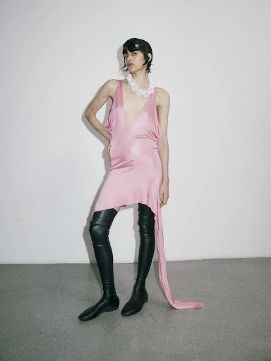 A young adult woman poses in a pink dress and thigh-high boots against a white background, creating a fashion-editorial aesthetic.