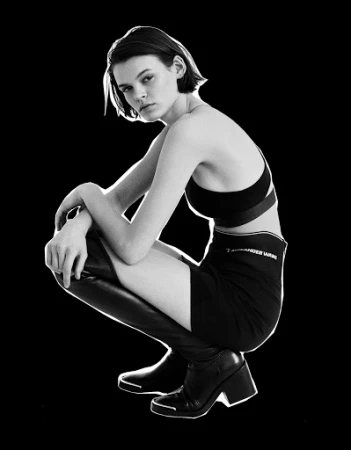Monochrome portrait of a young woman in a black sports bra, skirt, and high boots squatting against a stark black backdrop.