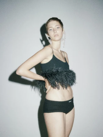 Young woman in black crop top with feather trim and black shorts poses in a studio setting with soft, diffused lighting.