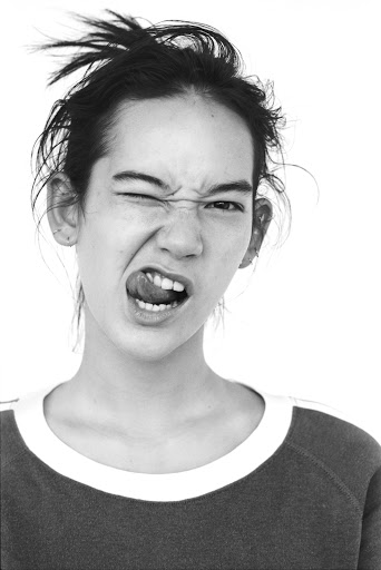 Black and white portrait of a young adult female with messy hair in a top, sticking out tongue, and winking in a photo studio.