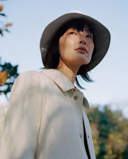 Asian woman in bucket hat and beige jacket gazes up in outdoor setting with dappled sunlight, creating a serene and fashionable portrait.