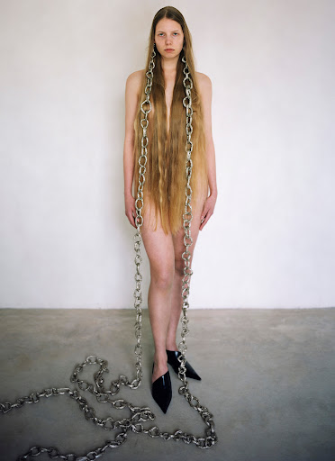Woman with long hair and chain necklaces poses against a white wall in black mules, creating a striking fashion image.