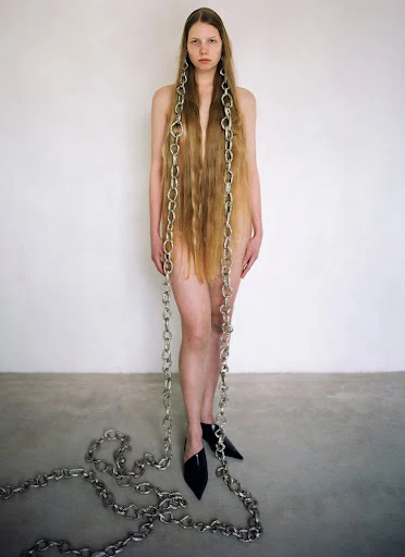 Woman with long hair and chain necklaces poses against a white wall in black mules, creating a striking fashion image.