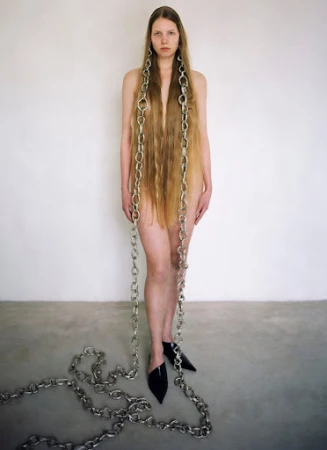Woman with long hair and chain necklaces poses against a white wall in black mules, creating a striking fashion image.