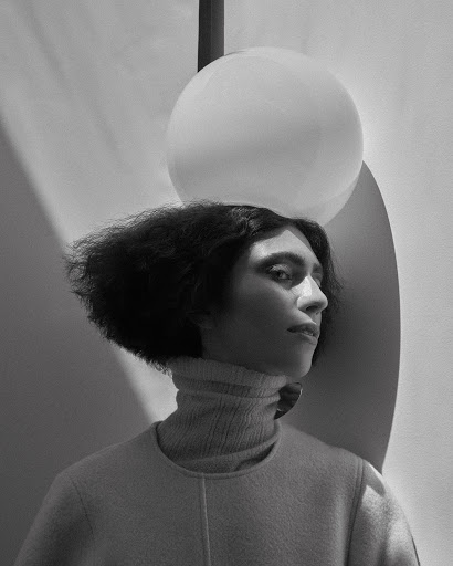 Black-and-white portrait of a person with dark, curly hair wearing a turtleneck sweater, posing in front of a spherical lamp and wall.