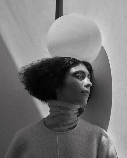 Black-and-white portrait of a person with dark, curly hair wearing a turtleneck sweater, posing in front of a spherical lamp and wall.