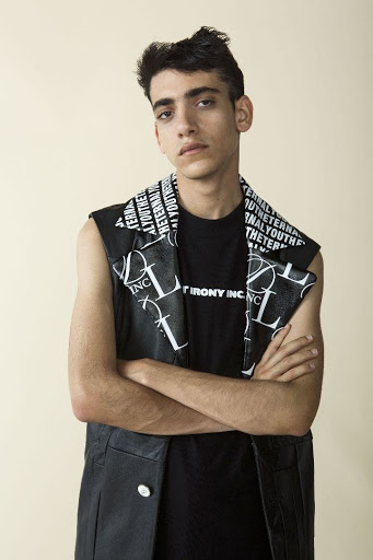 Portrait of a young adult male in a sleeveless jacket and patterned scarf, arms crossed, against a pale background. Fashion editorial.