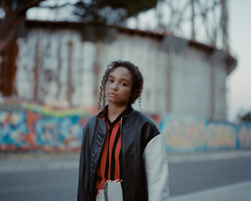 Portrait of a young Black woman with curly hair wearing a bomber jacket, standing outdoors in front of a graffiti-covered wall.