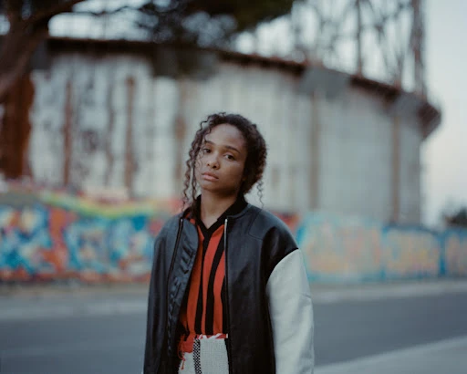 Portrait of a young Black woman with curly hair wearing a bomber jacket, standing outdoors in front of a graffiti-covered wall.
