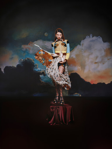 Woman stands on a pedestal wearing a colorful top and sheer skirt, posing against an artistic backdrop of clouds and night sky.