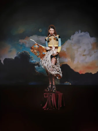 Woman stands on a pedestal wearing a colorful top and sheer skirt, posing against an artistic backdrop of clouds and night sky.