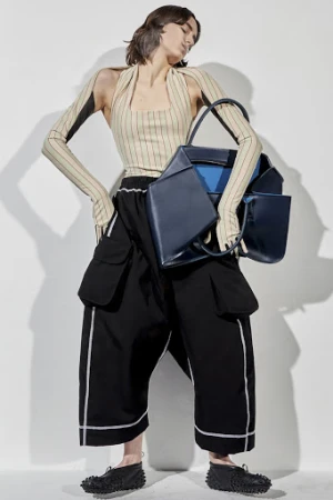 Model in striped halter top and black cargo pants holding a large dark blue handbag against a plain white studio backdrop.