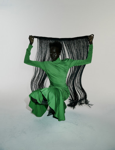 Model in a green dress crouches holding a hairpiece in a studio setting with a seamless white background, fashion editorial style.