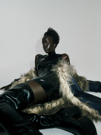 A Black fashion model in a black bodysuit and thigh-high boots poses against a white background, draped with a fur-trimmed coat.
