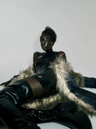 A Black fashion model in a black bodysuit and thigh-high boots poses against a white background, draped with a fur-trimmed coat.