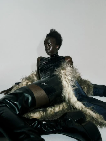A Black fashion model in a black bodysuit and thigh-high boots poses against a white background, draped with a fur-trimmed coat.