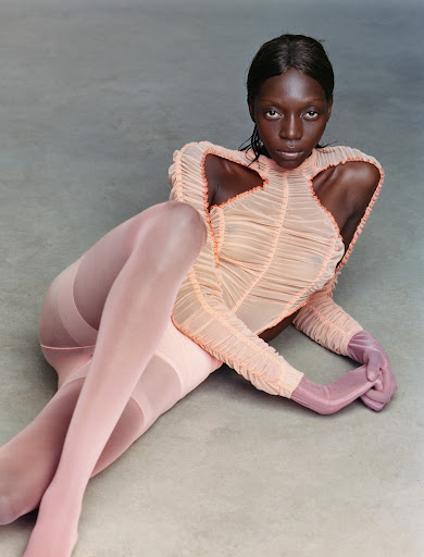 Fashion portrait of an African female model in a peach bodysuit and pink tights lying down in a studio against a light grey background.