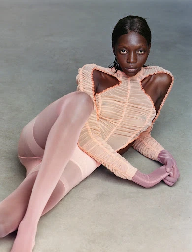 Fashion portrait of an African female model in a peach bodysuit and pink tights lying down in a studio against a light grey background.