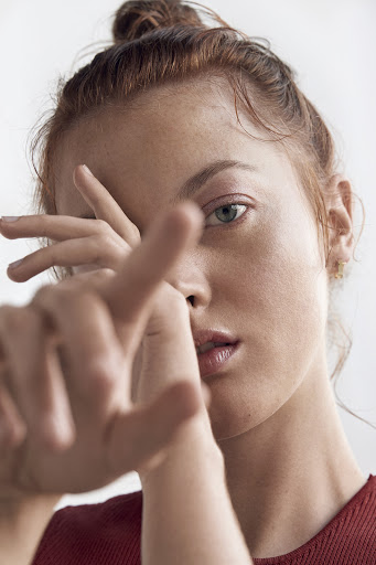 Close-up portrait of a red-haired woman with a hand raised in front of her face against a plain white backdrop in a studio setting.