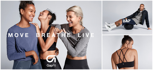 Three women laugh together in a GapFit advertisement. A man is balancing on a medicine ball and a sports bra has criss-cross straps.