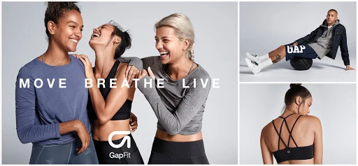 Three women laugh together in a GapFit advertisement. A man is balancing on a medicine ball and a sports bra has criss-cross straps.