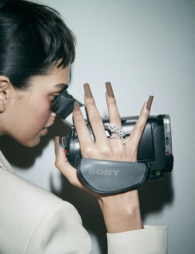 Adult woman with long brown nails filming with a Sony camcorder in a photo studio wearing a cream-colored blazer against a grey background.