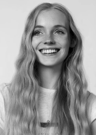 Portrait of a smiling young blonde woman with long wavy hair and freckles wearing a t-shirt against a plain white background.