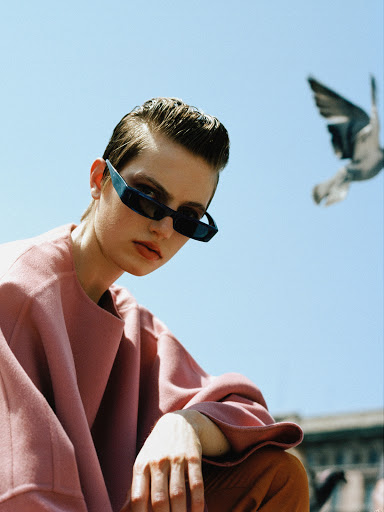 Eye-level shot of a short-haired model with sunglasses, a pink top, and orange pants posing against a clear blue sky with a pigeon.