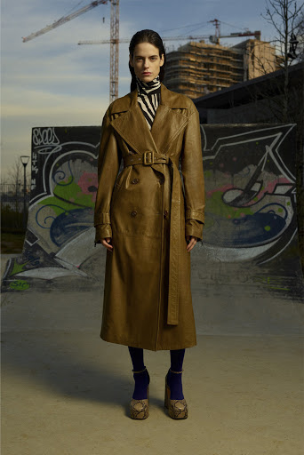 An adult female model wears a long, light brown trench coat, zebra print turtleneck, dark leggings, and snakeskin platform shoes in an urban setting.