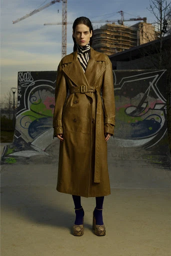 An adult female model wears a long, light brown trench coat, zebra print turtleneck, dark leggings, and snakeskin platform shoes in an urban setting.