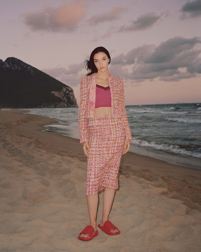 A young woman in a pink tweed suit stands on a sandy beach with ocean waves and a mountain visible against a colorful sky at dusk.