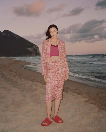 A young woman in a pink tweed suit stands on a sandy beach with ocean waves and a mountain visible against a colorful sky at dusk.