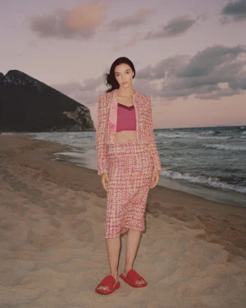 A young woman in a pink tweed suit stands on a sandy beach with ocean waves and a mountain visible against a colorful sky at dusk.