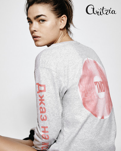 A young woman poses indoors in a grey long-sleeve shirt with a graphic. Fashion and beauty editorial content for a creative platform.