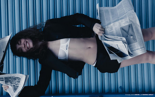 Woman in bra top and blazer lies on blue corrugated surface holding newspapers. Fashion editorial with beauty and minimal clothing.