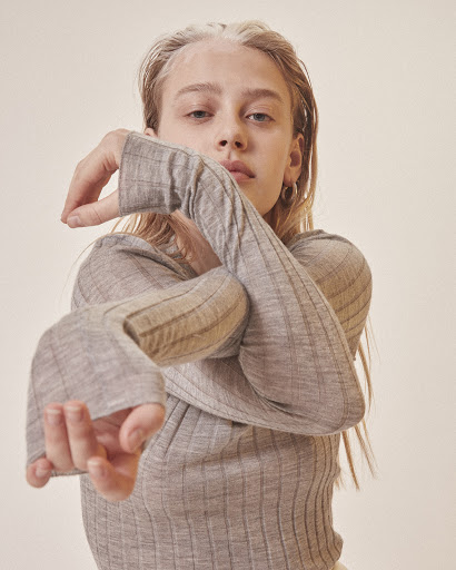 Blonde woman in ribbed grey top with long sleeves poses in a studio setting with neutral backdrop in a portrait orientation.